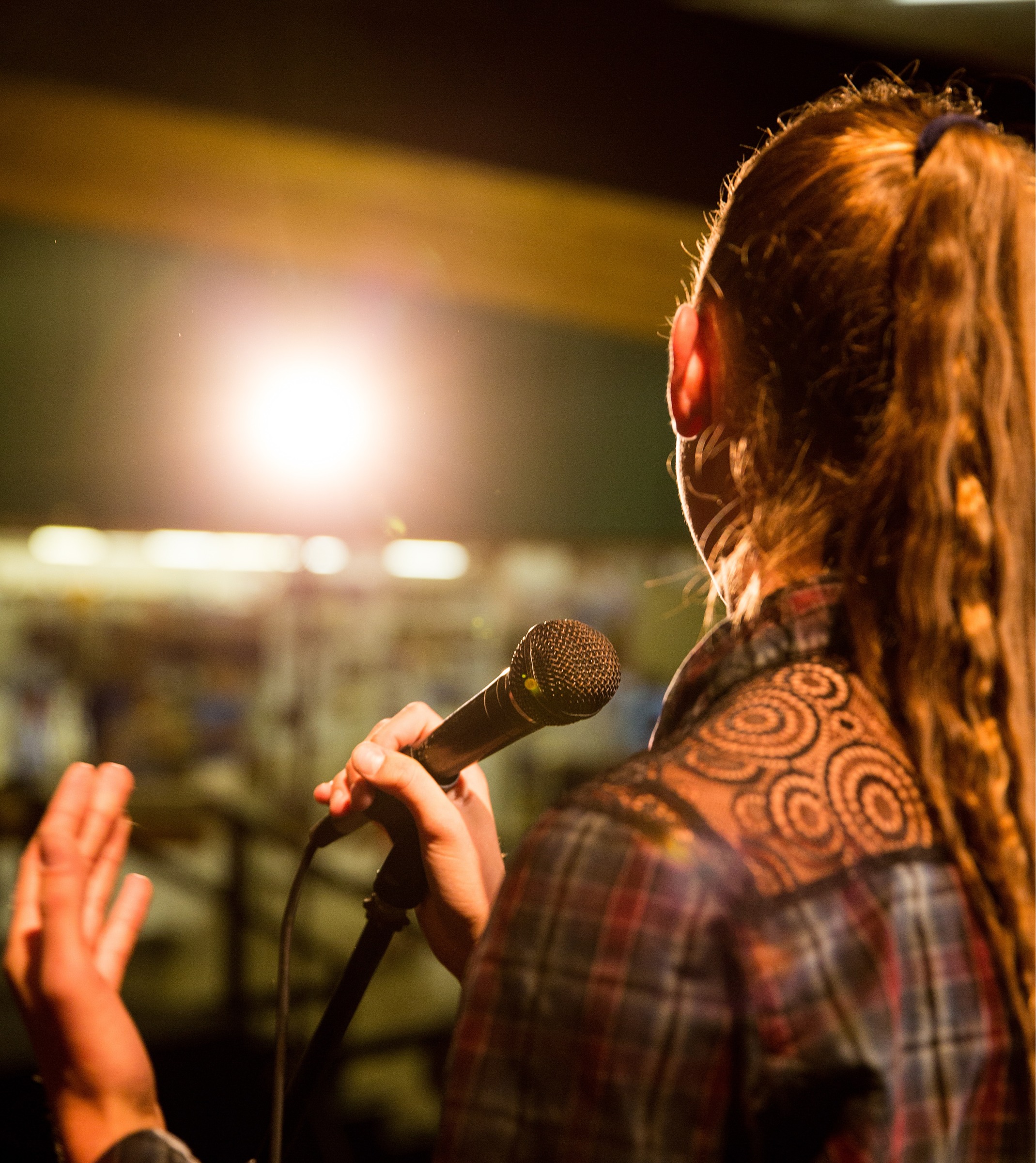 A young person with wavy, light brown hair in a ponytail has their back to the camera. They are holding a microphone.