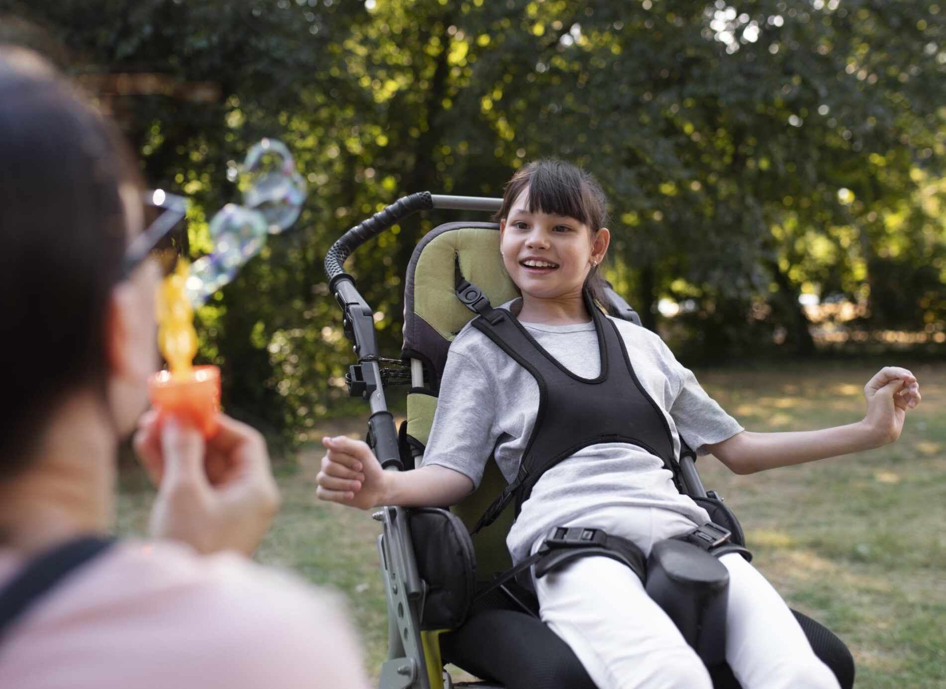 A young girl in an adaptive chair grins as another person blows bubbles towards her in a park.