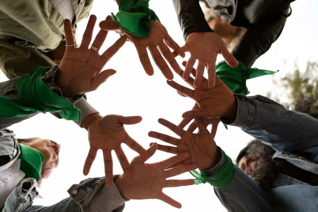 A multiracial group of young people hold out their hands over the camera, which points up to the sky. They wear green bandanas around their wrists.