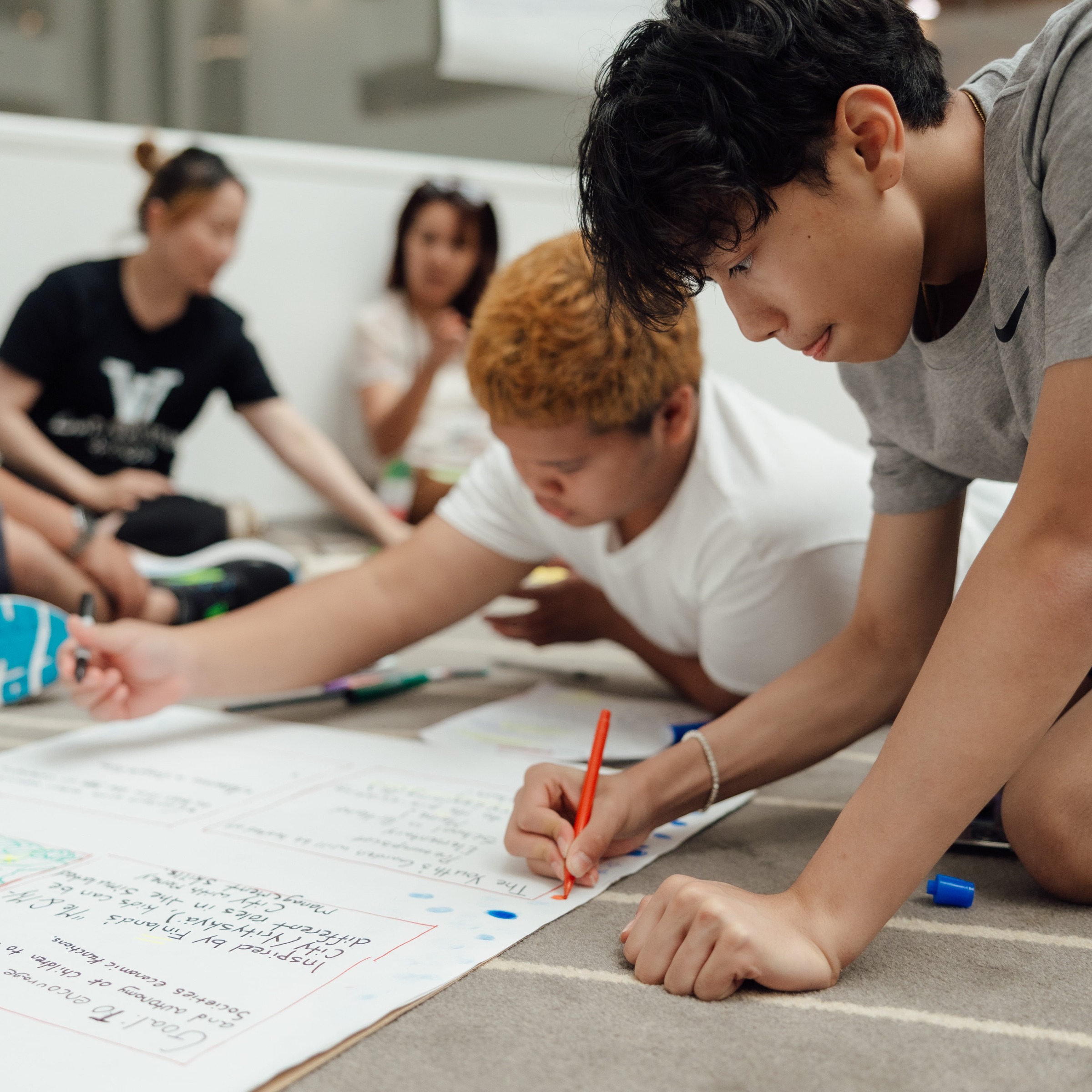 Two young Asian American men write on a poster board on the floor.