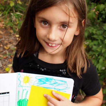 A girl with pale skin and long brown hair holds up a drawing of a landscape. She is outdoors.