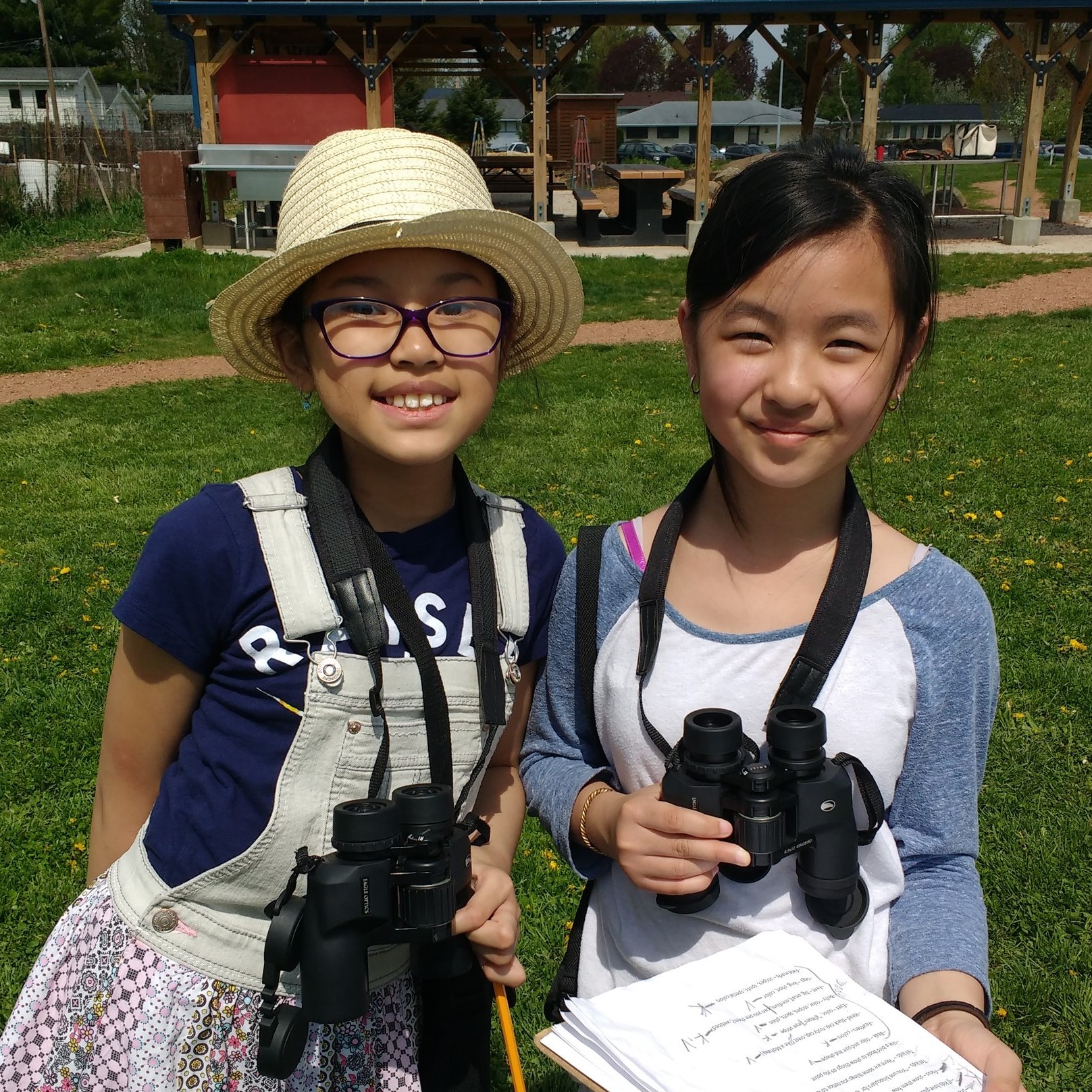 Two Asian American girls stand outdoors smiling and holding birding binoculars.