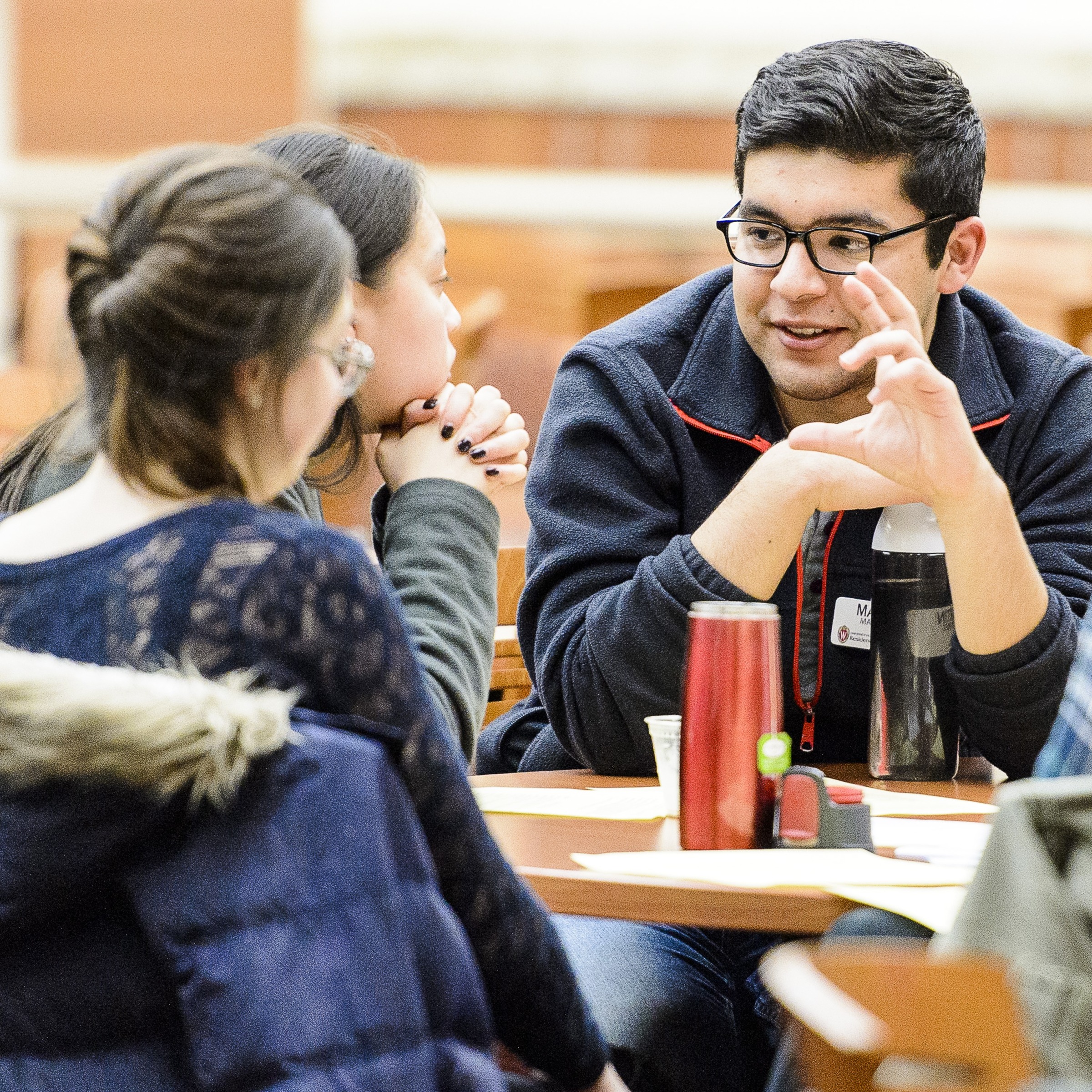 A man with dark hair and pale skin sits at a table with two young women, who also have straight, dark hair. He makes eye contact and smiles while talking.